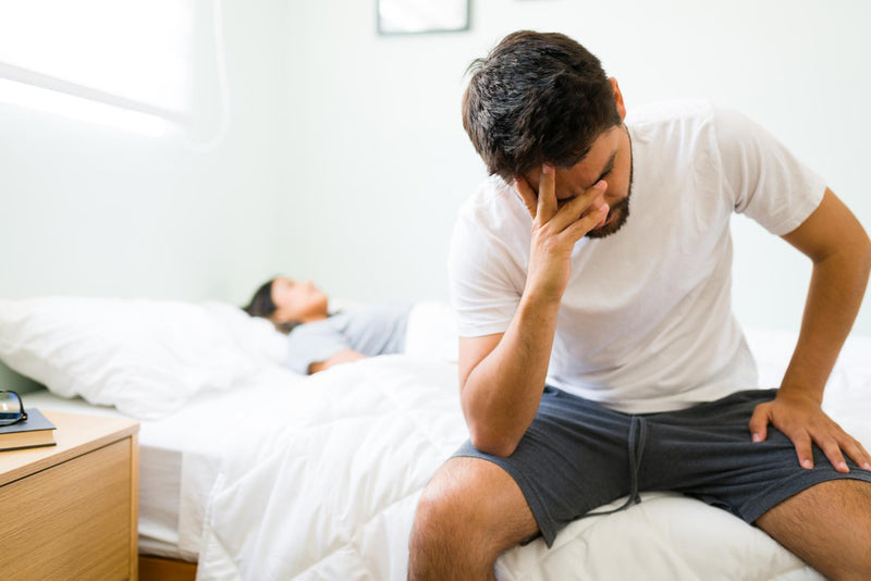 Man sitting on a bed with a distressed expression, another person lying in the background.
